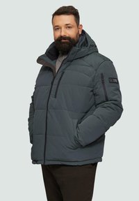 Donkergroene pufferjack met een afneembare capuchon. Voorzien van ritszakken, horizontale stiksels en een logo patch op de linkerschouder.