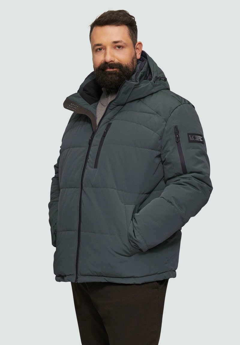 Donkergroene pufferjack met een afneembare capuchon. Voorzien van ritszakken, horizontale stiksels en een logo patch op de linkerschouder.