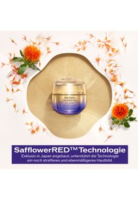 Shiseido Vital Perfection -voide lasipurkissa, jossa on kirkas kansi ja kullasta violettiin oleva liukuvärjäys. Ympärillä saflorinkukkien terälehtiä ja lehtiä.