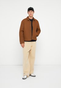 Brown zip-up jacket with a collar, worn over a dark shirt, paired with light beige pants and black sneakers, accentuated by a black cap.