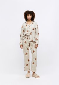 Beige pyjama set met een overhemd met knopen en broek met een trekkoord, voorzien van een teddybeerpatroon. Zachte textuur en een ontspannen pasvorm, gecombineerd met donzige slippers.