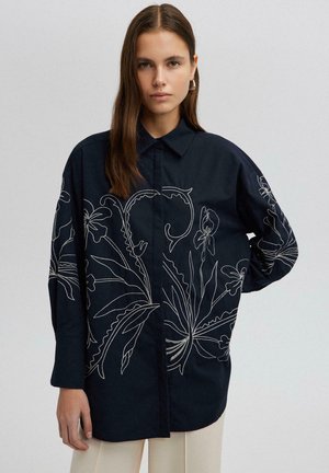 Camicia blu navy con maniche lunghe e colletto a punta, con ricami floreali bianchi sul davanti e sulle maniche.