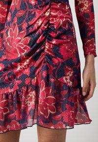 Robe à motif floral rouge et bleu marine avec des détails froncés et un ourlet à volants portée par un mannequin avec une main visible.