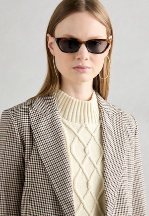 Schildpatt-Cat-Eye-Sonnenbrille, cremefarbener Zopfstrickpullover und ein brauner Karoblazer mit auffälligen Revers und strukturiertem Schnitt.