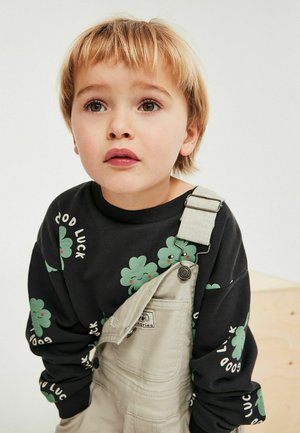 Junges Kind mit blonden Haaren, trägt schwarzen Pullover mit grünen Kleeblättern und der Aufschrift "Viel Glück", beige Latzhose, schaut leicht nach oben mit neutralem Gesichtsausdruck.