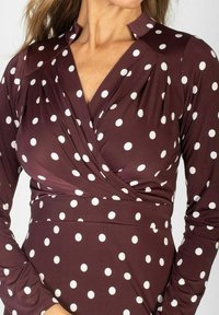Kobieta w bordowej sukience zapinanej z białymi kropkami i długimi rękawami, pokazująca górną część ciała i dekolt.