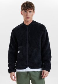 Svart, fluffig fleecejacka med dragkedja fram, ståkrage och sidoficka. Har ribbade ärmslut och en avslappnad passform.