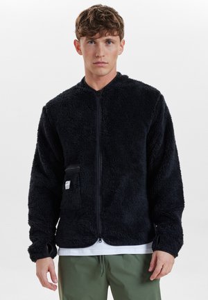 Chaqueta de forro polar negra y suave con cremallera frontal, cuello alzado y bolsillos laterales. Presenta puños de canalé y un ajuste relajado.