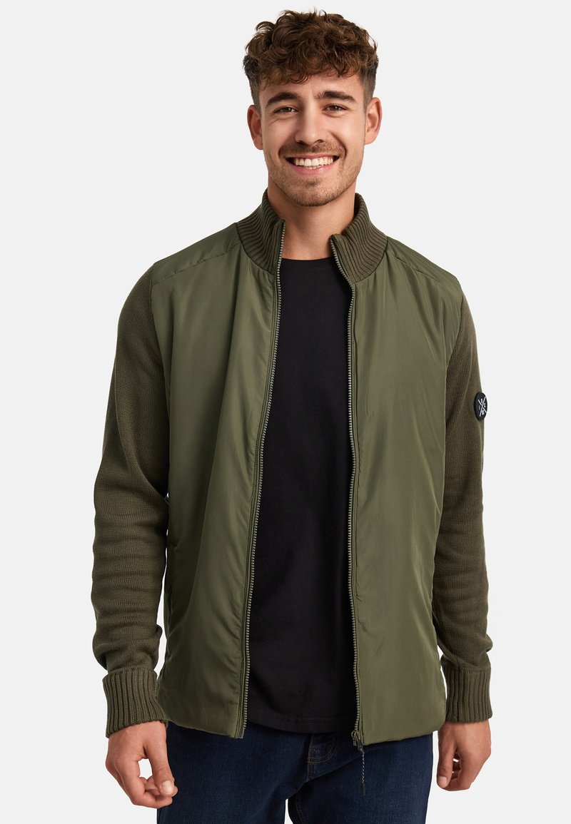 Chaqueta verde olivo con un diseño de punto, que presenta un cuello alto, cremallera completa y puños acanalados. Camisa negra debajo, jeans de mezclilla.