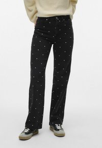 Sorte denimjeans med hvite prikkar, høg midje, vida bein, med knapp og glidelås. Brukt sammen med svart og grå sneakers.