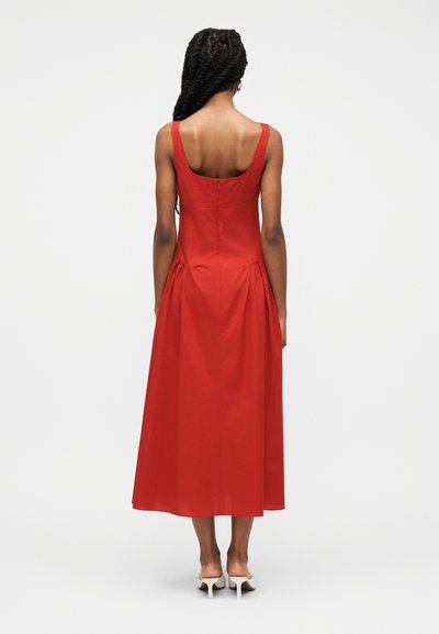 Γυναίκα με πλεξούδες, που φοράει μια κόκκινη midi αμάνικη φόρεμα και λευκές γόβες, στέκεται γυρισμένη με την πλάτη σε έναν απλό φόντο.