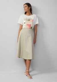 Witte cropped t-shirt met een gekleurd abstract ontwerp en gouden accenten, gecombineerd met een beige, knielange, A-lijn rok. Strappy witte sandalen.