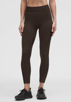 Vrouw die donkerbruine legging met hoge taille en zwarte sportschoenen draagt, staand tegen een effen lichte achtergrond.