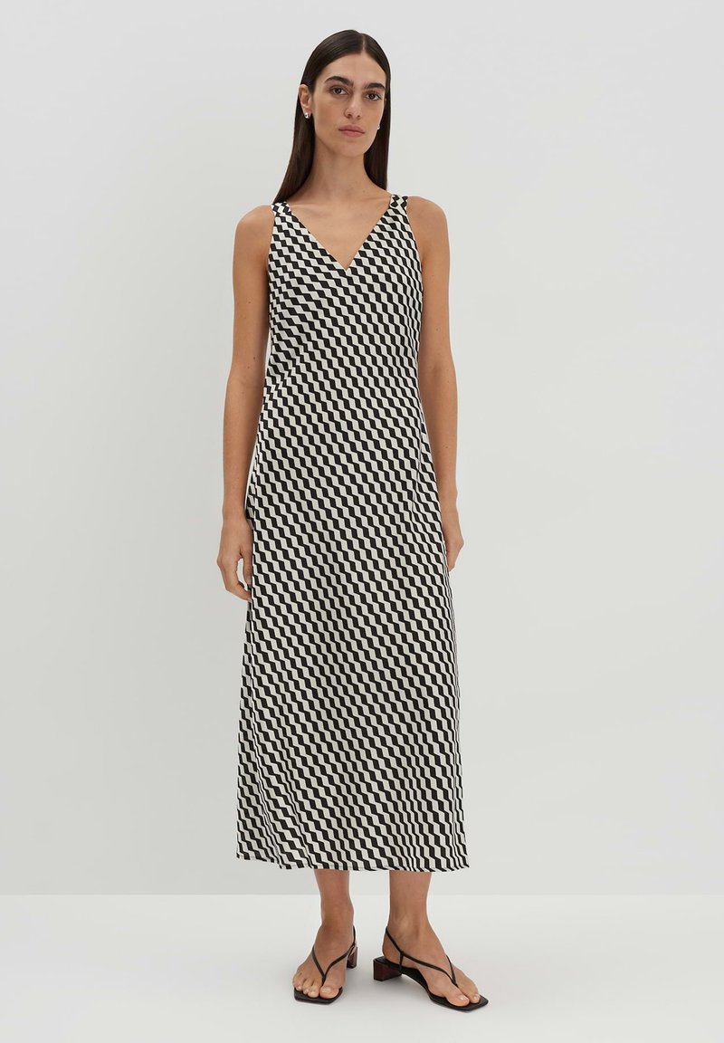 Ärmelloses, V-Ausschnitt-Kleid im schwarz-weißen Hahnentrittmuster. Fließende Silhouette, knielang, weicher Stoff, kombiniert mit minimalen Absatzsandalen.