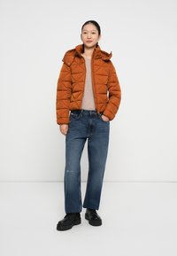 Brun pufferjakke med diamantkviltet tekstur, hætte og lynlås lukning. Båret over en lys sweater og parret med brede blå jeans og sorte støvler.