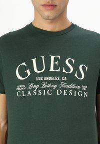 Tumši zaļš kokvilnas T-krekls ar baltu tekstu. Dizainā iekļauti uzraksti "GUESS," "Los Angeles, CA," un "Klasicisks dizains".