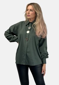 Ciemnozielona koszula zapinana na guziki, wykonana z gładkiego materiału, z dużymi srebrnymi guzikami i długimi rękawami; zestawiona z czarnymi skórzanymi spodniami.