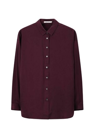 Femme aux cheveux courts portant des boucles d'oreilles en or, une chemise boutonnée violet foncé et un pantalon rose texturé et ample, debout devant un fond beige.