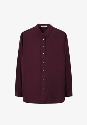 Vrouw met kort haar, die gouden oorbellen draagt, een donkerpaarse blouse met knopen en losse roze textuurbroek, staand tegen een beige achtergrond.