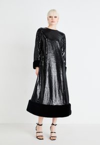Dolga črna obleka s sequinom z sijočo teksturo, ki ima dolge rokave in črn krzneni rob. Nosi se z črnimi sandalnimi petami.