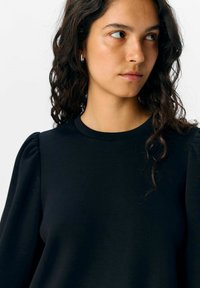 Haut noir à manches longues avec encolure arrondie, doté de manches bouffantes. Fabriqué dans un tissu lisse avec un léger éclat. Aucun motif ou accent notable.