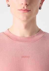 Sudadera rosa con textura acanalada y cuello redondo. Presenta un pequeño logo emblema "HUGO" en el pecho.