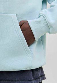 Sudadera azul claro con bolsillo tipo canguro, puños acanalados y tela contrastante oscura en la parte inferior. Textura suave y diseño minimalista.