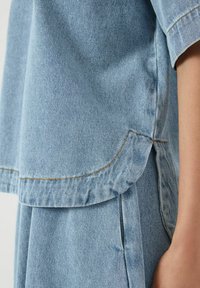 Top in denim azzurro chiaro con maniche corte, caratterizzato da un orlo arrotondato e cuciture arancioni sottili. Il tessuto ha una finitura liscia e strutturata.