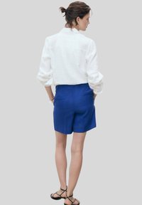 Biela ľanová košeľa s vyhrnutými rukávmi v kombinácii s modrými šortkami s vysokým pásom, ktoré majú zadné vrecká a voľný strih.