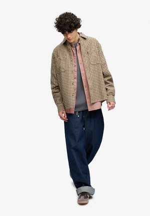 Ung person med krøllet hår iført lagdelte ternede og strikkede skjorter, løse mørke jeans og sneakers, står mod en hvid baggrund.