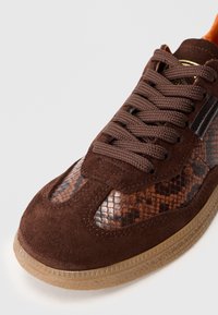 Barna velúr sportcipő kígyóbőr minta panellel és barna fűzőkkel. Gumitalpa van, amely textúrázott mintázattal rendelkezik, és kerek orrú kialakítással rendelkezik.