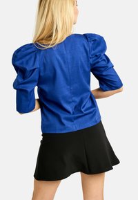 Modrá blúzka s nafúknutými rukávmi a textúrovaným povrchom, kombinovaná s čiernou rozšírenou sukňou. Blúzka má priliehavý zadný diel a krátke rukávy.
