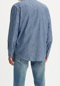 Camicia in chambray blu a maniche lunghe con yoke curvo, polsini con bottoni e fondo arrotondato, abbinata a jeans in denim chiaro.