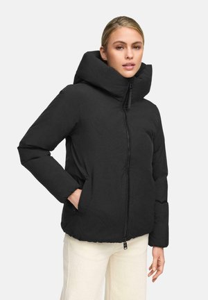 Woman wearing a black puffer jacket with a high collar and zipper, hands in pockets, paired with light-colored pants, against a plain background.
