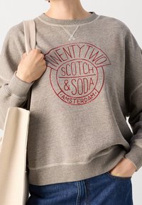 Sudadera gris con texto gráfico en rojo. Presenta un escote redondo, puños acanalados y un ajuste relajado. Combinada con un bolso tote beige.