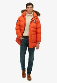 Puffjakke i lys orange, med en pelsforet hætte, tonede syninger, et patch-logo og lukket med knapper og lynlåse.
