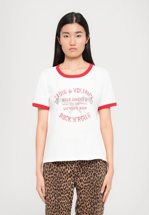 T-shirt blanc à manches courtes avec bord rouge, affichant un texte pailleté : "Zadig & Voltaire Rock'n'Roll." Porté avec un pantalon imprimé léopard marron.