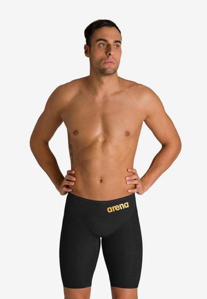 Muž bez trička v čiernych plaveckých šortkách Arena po kolená so zlatým logom, stojaci s rukami v bok na jednoliatom pozadí.