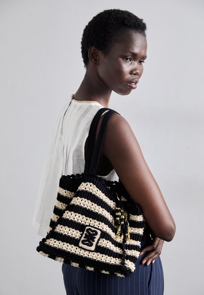 Sac à main tissé noir et beige à rayures avec un accent de pompon et un patch logo. Il dispose de poignées tressées et d'une forme structurée.