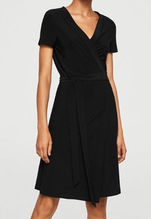 Frau trägt ein schwarzes Wickelkleid knielanger Länge mit kurzen Ärmeln und einem gebundenen Taillengürtel vor einem einfarbigen Hintergrund.