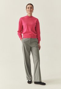 Pull rose à manches longues et col montant, associé à un pantalon gris clair à jambes larges et des chaussures noires avec des lacets beiges. Fond uni.