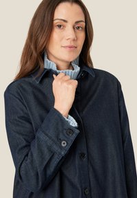 Mørk denim skjortejakke med krave, sorte knapper og en lyseblå ribbet tætsiddende sweater under. Tekstureret stof, afslappet pasform.