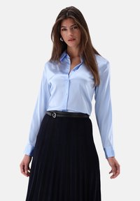 Helltürkise Satinbluse mit einem Button-Down-Kragen, kombiniert mit einem schwarzen Faltenrock und einem schmalen schwarzen Gürtel. Das Outfit hat lange Ärmel.
