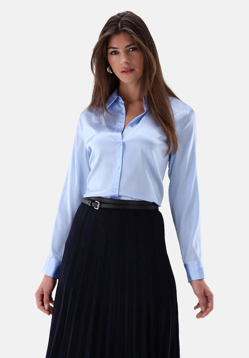Helltürkise Satinbluse mit einem Button-Down-Kragen, kombiniert mit einem schwarzen Faltenrock und einem schmalen schwarzen Gürtel. Das Outfit hat lange Ärmel.