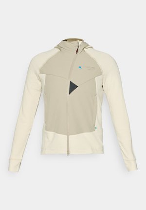 Bež in krema zip-up jakna z teksturovanim vzorcem, ki ima kapuco, geometrijske črne poudarke in logo na prsih.