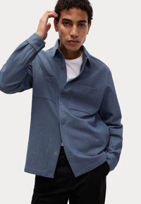 Modrá košile na knoflíky s plochým límcem, dvěma náprsními kapsami, texturovanou tkaninou a pohodlným střihem, nošená přes bílé tričko.