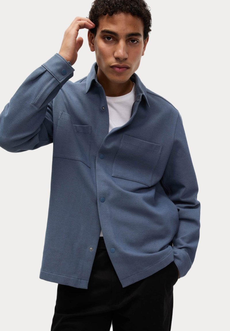 Modrá košile na knoflíky s plochým límcem, dvěma náprsními kapsami, texturovanou tkaninou a pohodlným střihem, nošená přes bílé tričko.