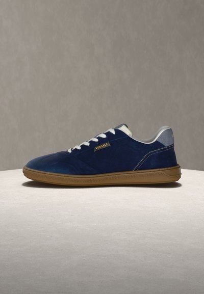 Marineblaue Wildleder-Sneaker in niedrigem Schnitt mit weißen Schnürsenkeln, beigefarbener Gummisohle und kleinem goldenen Versace-Logo an der Seite.