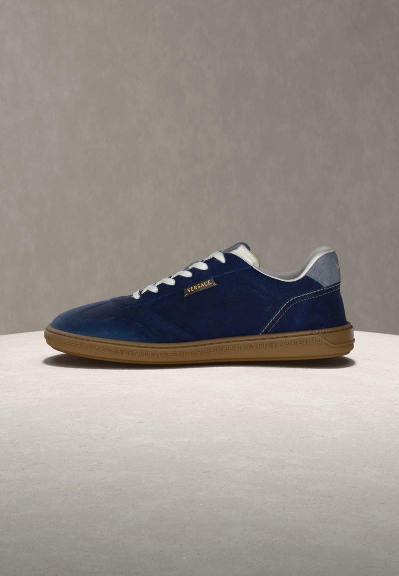 Sneaker blu navy in camoscio con tomaia bassa, lacci bianchi, suola in gomma color cuoio e piccolo logo Versace dorato sul lato.