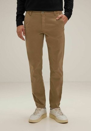 Beige Chinos aus Baumwolle mit glatter Textur, featuring seitliche Taschen und einen vorderen Knopfverschluss. Kombiniert mit weißen Sneakers.
